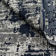 A close-up of a woven fabric with an abstract distressed pattern in navy blue, charcoal gray, and off-white, with a folded corner showing the texture and frayed edge.