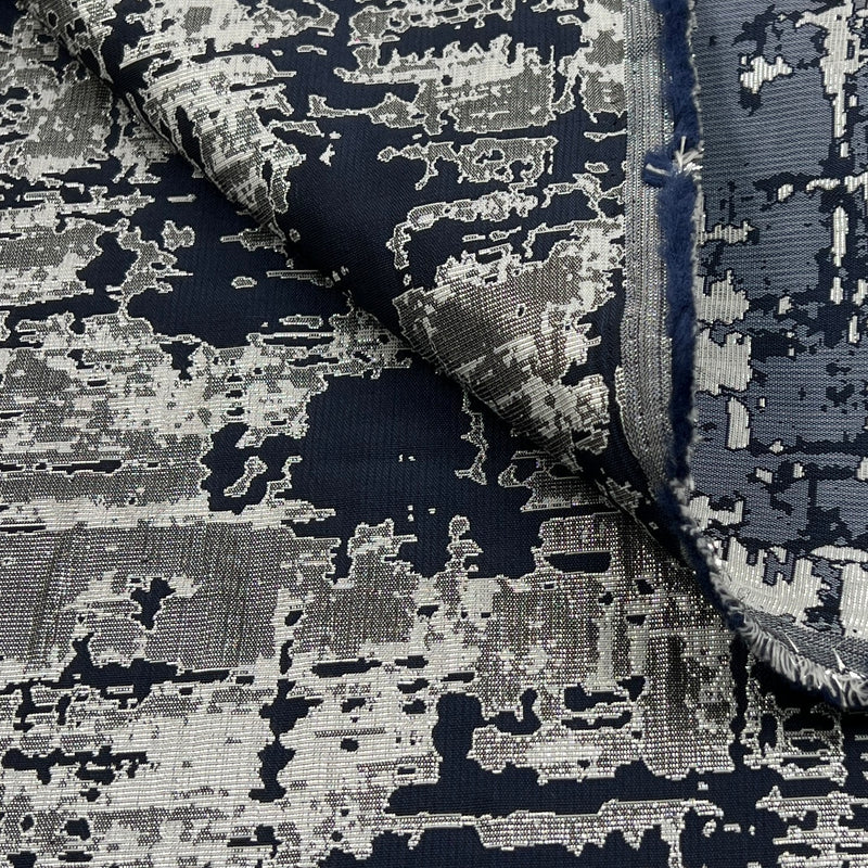 A close-up of a woven fabric with an abstract distressed pattern in navy blue, charcoal gray, and off-white, with a folded corner showing the texture and frayed edge.