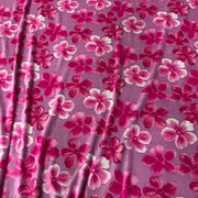 A close-up of a purple fabric with a repeating floral pattern of pink and magenta flowers with white highlights.