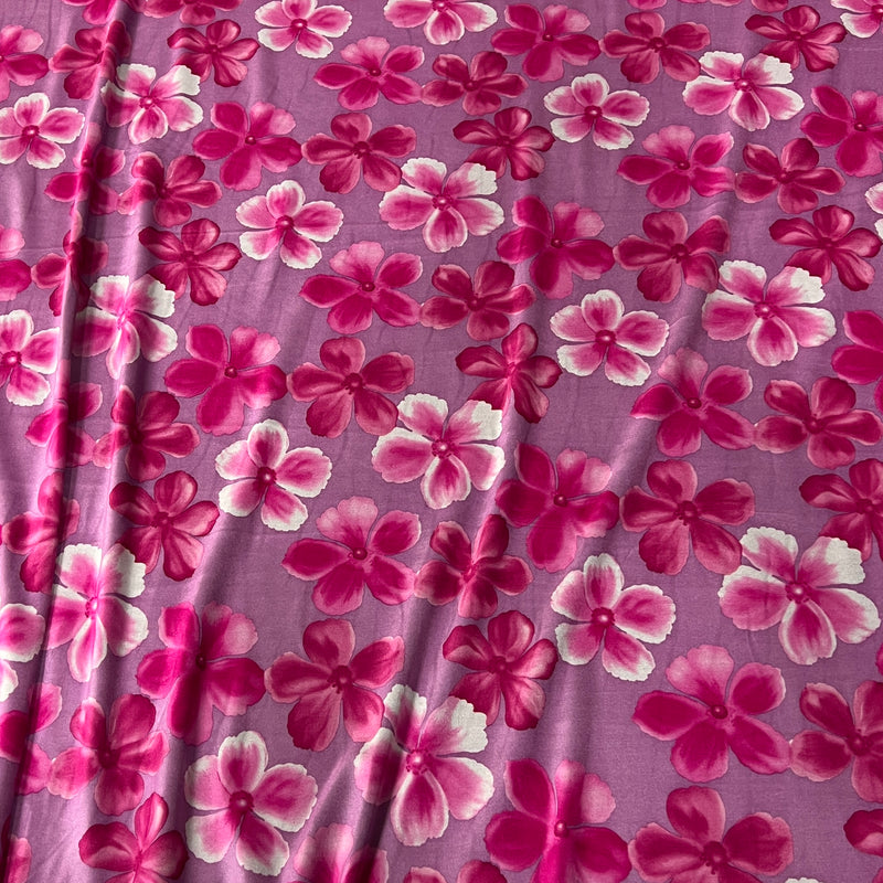 A close-up of a purple fabric with a repeating floral pattern of pink and magenta flowers with white highlights.