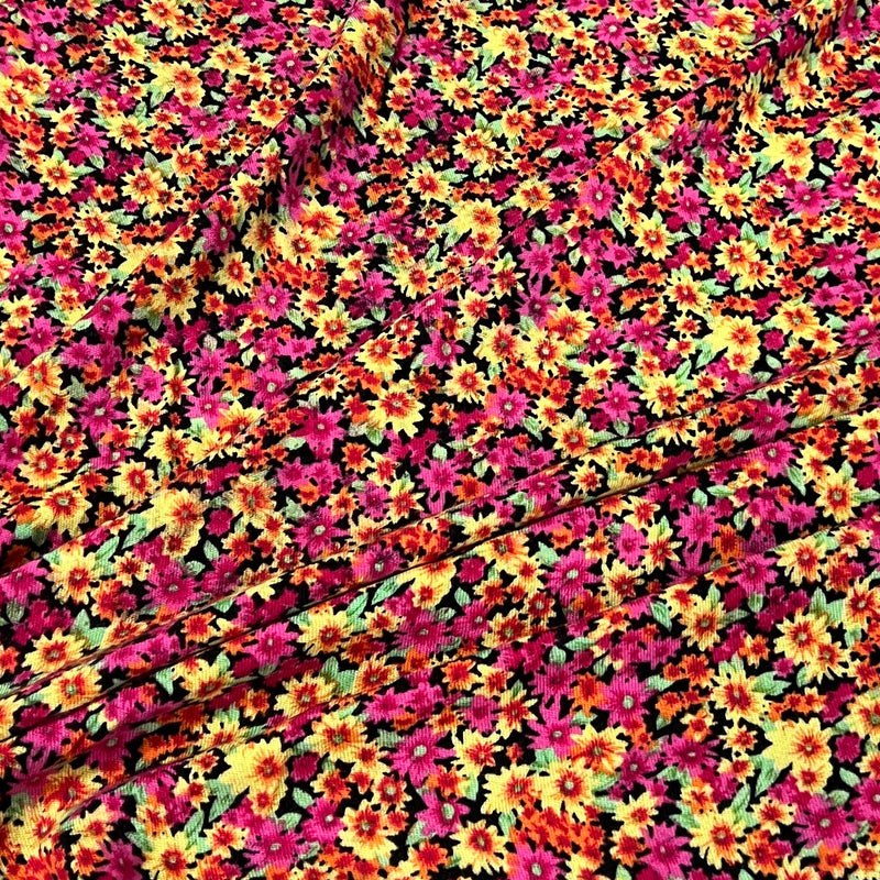 A close-up of a vibrant floral fabric featuring a dense pattern of small daisy-like flowers in magenta pink, bright yellow, orange, and green on a black background.