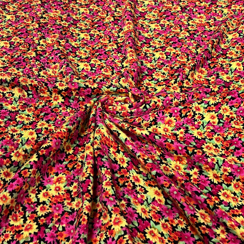 A close-up of a draped fabric with a dense, all-over floral print featuring bright pink, yellow, orange, and green flowers on a black background.