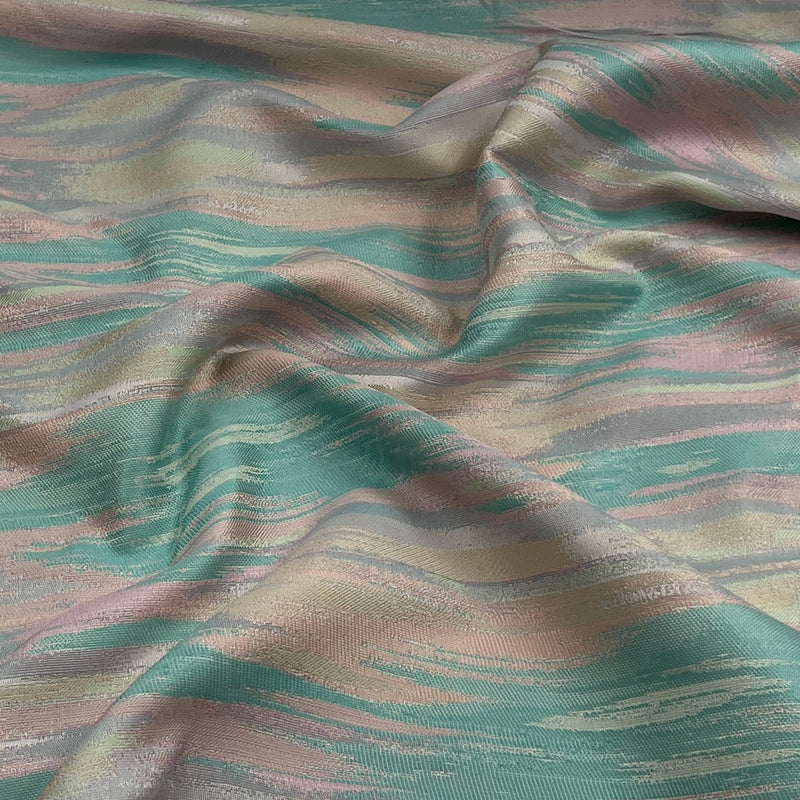 A close-up of draped fabric with a shimmering abstract pattern in teal green, dusty pink, and warm beige tones.