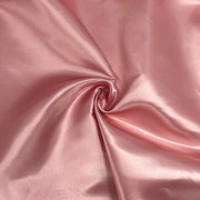 A close-up of draped satin fabric in a soft dusty pink color, showing a glossy sheen and smooth texture with a central swirl fold.