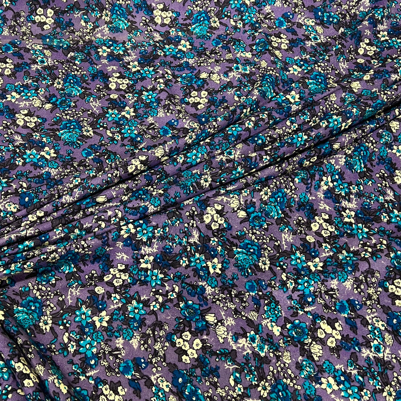 Purple floral fabric with a dense pattern of blue, teal, and cream flowers, draped diagonally across the frame.