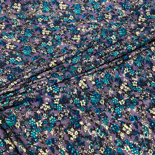 Purple floral fabric with a dense pattern of blue, teal, and cream flowers, draped diagonally across the frame.