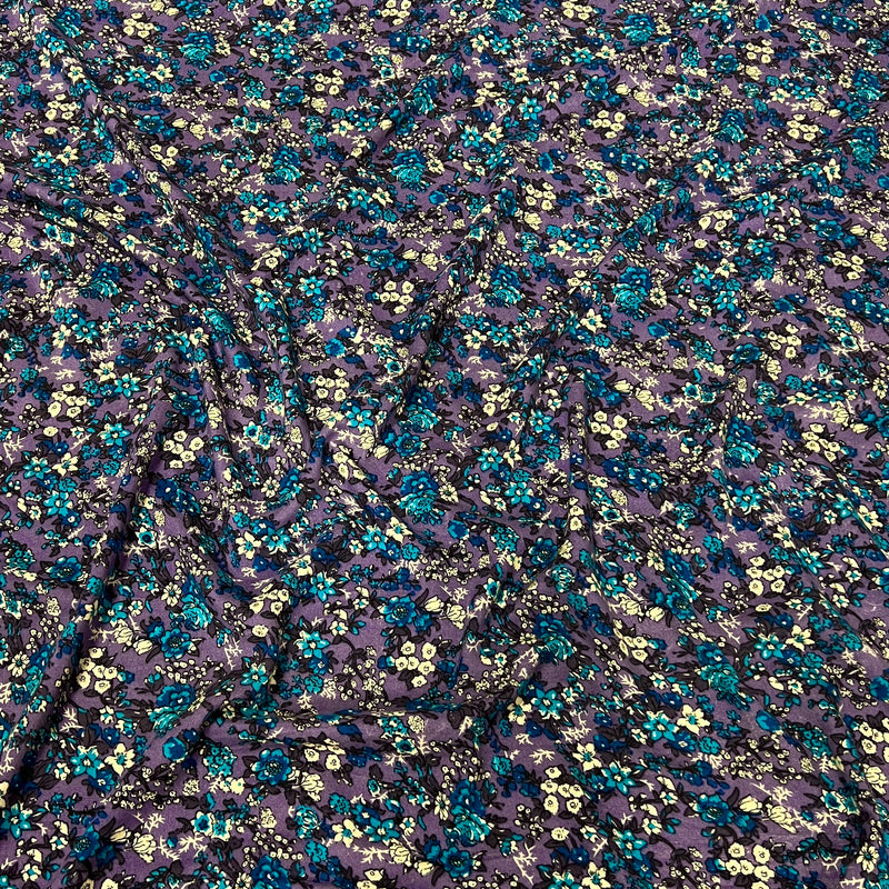 A close-up of a sheer purple mesh fabric with an all-over dense floral print in teal blue, yellow, and black.