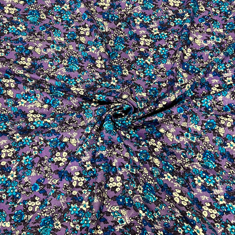 Purple floral fabric with a dense pattern of blue, teal, and cream flowers, gathered in the center to show its drape and texture.