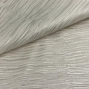 A close-up of textured fabric with a wavy, ribbed pattern in a light warm gray or champagne tone, folded over in the upper left corner.