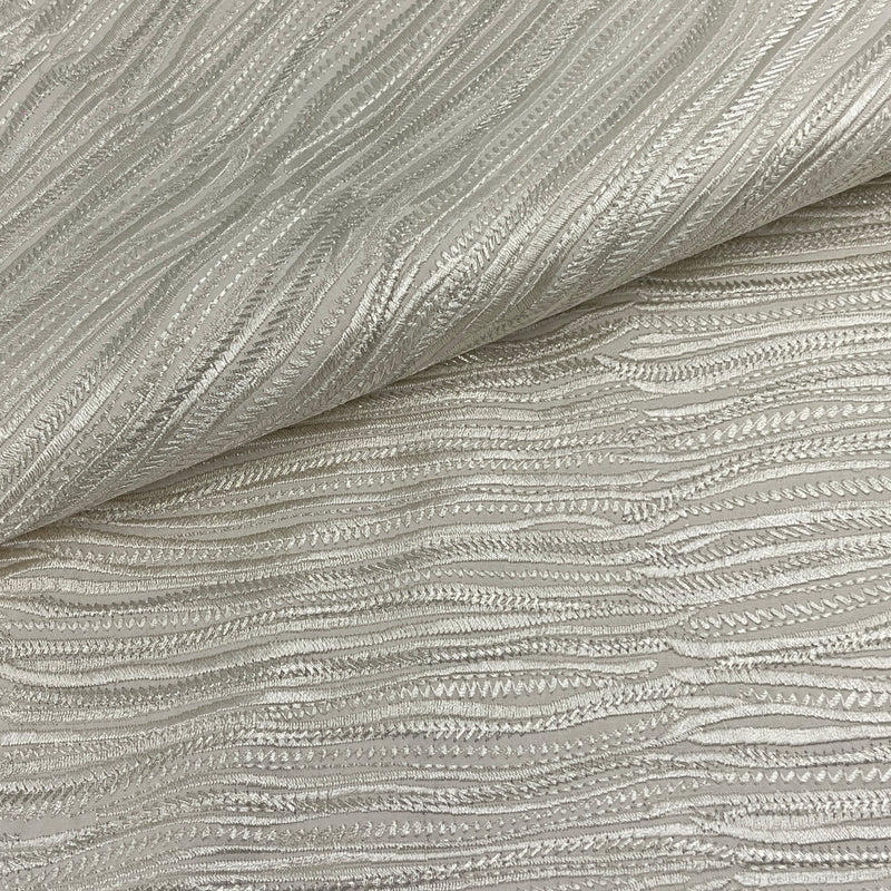 A close-up of textured fabric with a wavy, ribbed pattern in a light warm gray or champagne tone, folded over in the upper left corner.