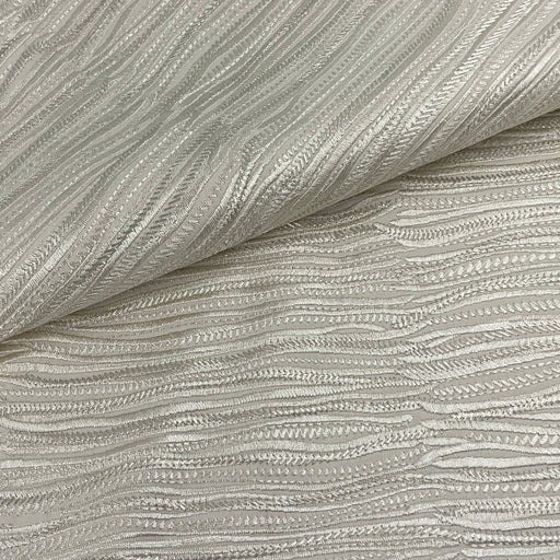 A close-up of textured fabric with a wavy, ribbed pattern in a light warm gray or champagne tone, folded over in the upper left corner.