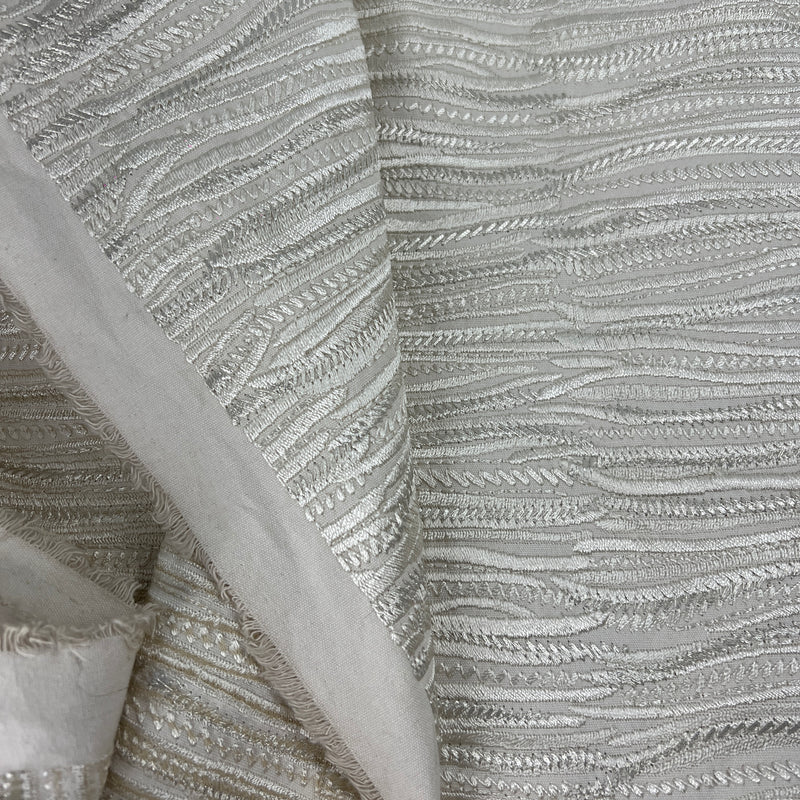 A close-up of textured fabric with horizontal wavy stripes in metallic silver and light gray, with a folded section showing the material's soft edge.