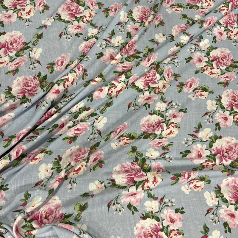 Light blue-gray floral fabric with a repeating print of pink and white roses, green leaves, and small yellow accents, shown draped with soft folds.