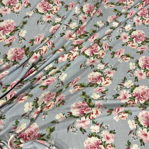 Light blue-gray floral fabric with a repeating print of pink and white roses, green leaves, and small yellow accents, shown draped with soft folds.
