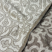 A close-up of a woven fabric with an intricate floral and scroll pattern in beige and taupe tones, showing the texture and a folded edge.