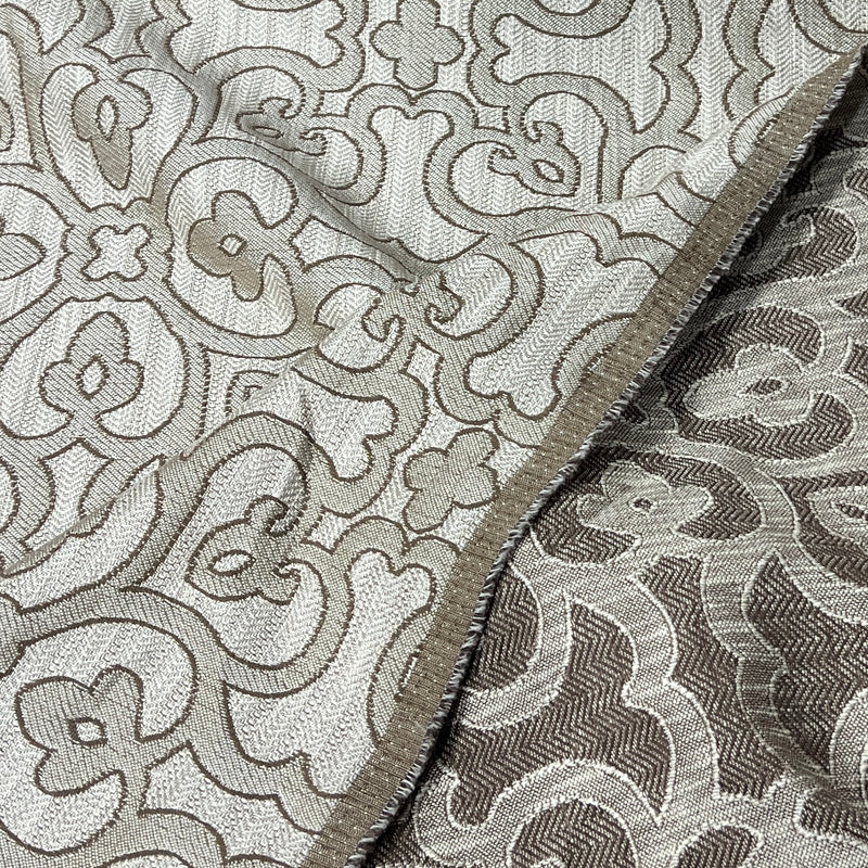 A close-up of a woven fabric with an intricate floral and scroll pattern in beige and taupe tones, showing the texture and a folded edge.