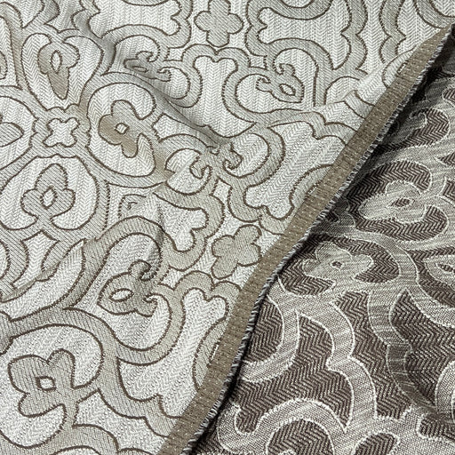 A close-up of a woven fabric with an intricate floral and scroll pattern in beige and taupe tones, showing the texture and a folded edge.