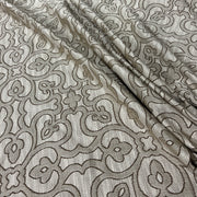 A close-up of a woven fabric with an intricate floral and scroll pattern in taupe and light beige, showing the texture and drape of the material.