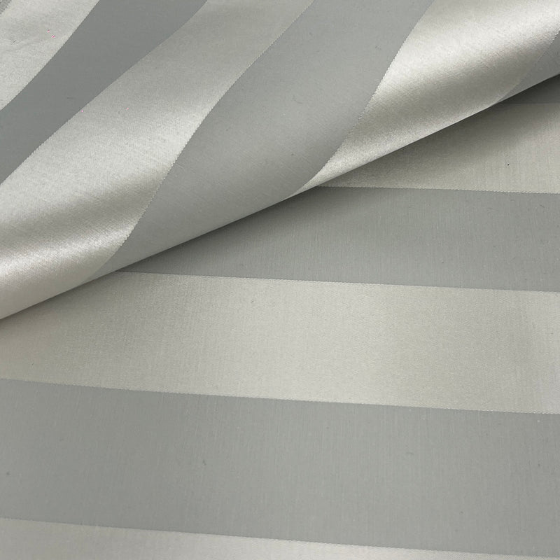 A close-up of a striped fabric or wallpaper pattern with alternating wide and narrow metallic-looking stripes in silver and light gray, shown with a folded corner revealing the texture.