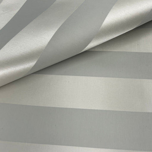 A close-up of a striped fabric or wallpaper pattern with alternating wide and narrow metallic-looking stripes in silver and light gray, shown with a folded corner revealing the texture.