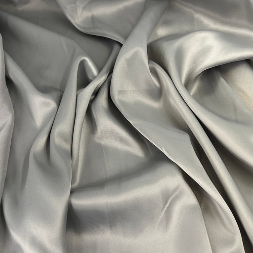 A close-up of draped satin fabric in a light silver-gray color, showing its smooth texture and sheen.