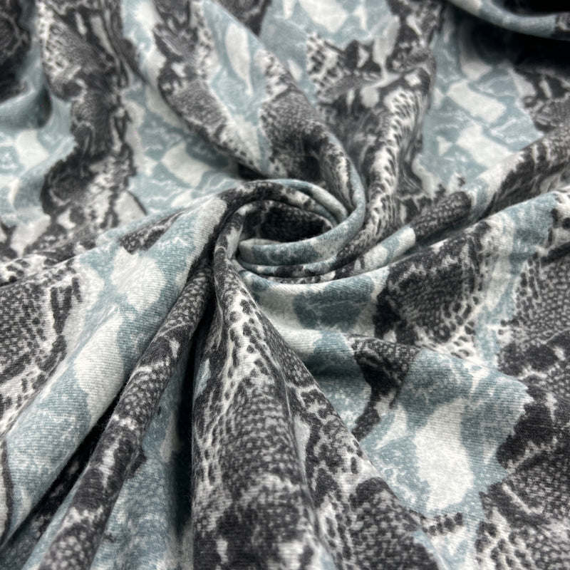 A close-up of draped fabric with a snakeskin pattern in muted blue-gray, charcoal gray, and off-white, gathered in the center to show the material texture and pattern repeat.