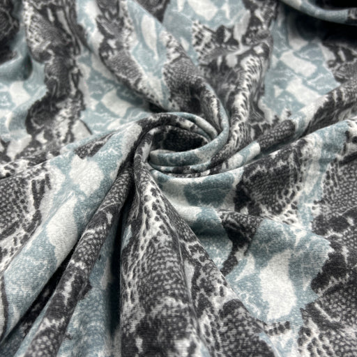 A close-up of draped fabric with a snakeskin pattern in muted blue-gray, charcoal gray, and off-white, gathered in the center to show the material texture and pattern repeat.