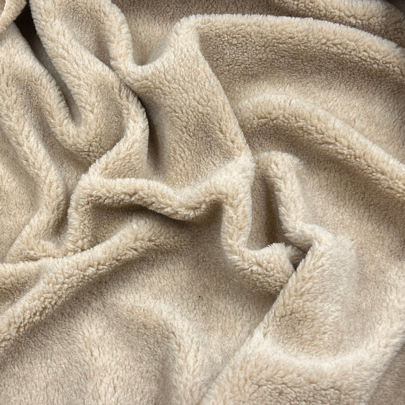 A close-up of soft, plush fleece fabric in a warm light beige or oatmeal color, shown with gentle folds and texture.