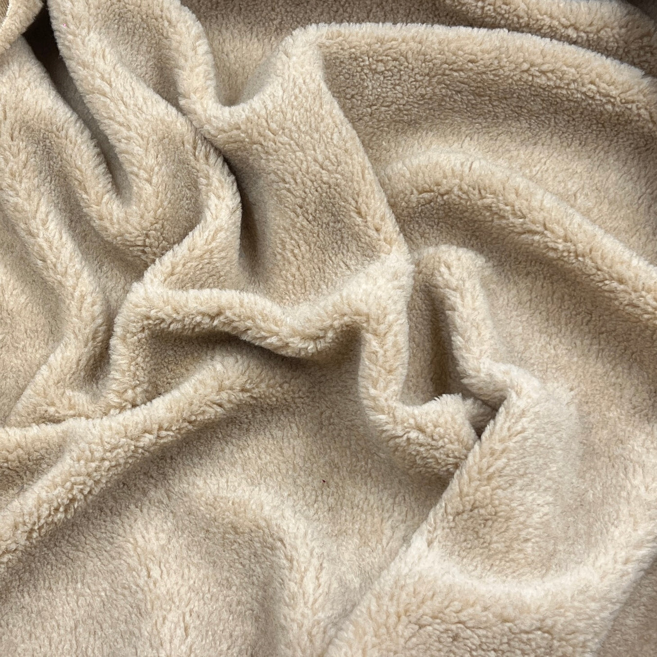 A close-up of soft, plush fleece fabric in a warm light beige or oatmeal color, shown with gentle folds and texture.