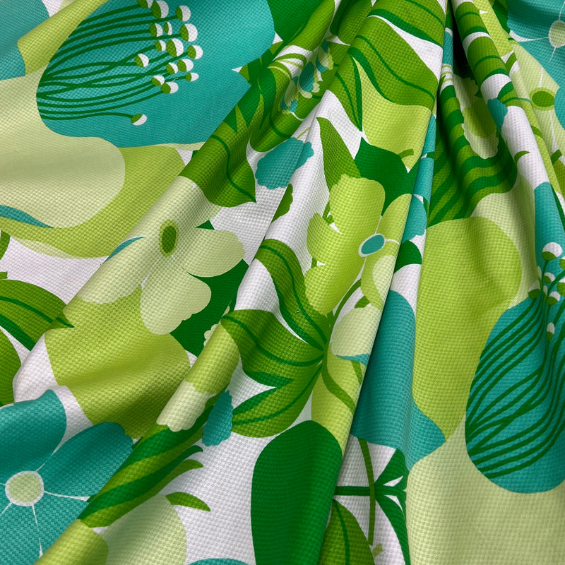 A close-up of draped fabric with a bold retro floral print in green, teal, and light yellow on a white background.