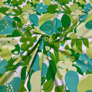 A close-up of a fabric with a retro 1970s floral pattern featuring green, teal, and light yellow flowers on a white background, gathered in the center to show its drape and texture.