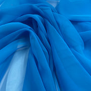 A close-up of draped bright blue sheer fabric with soft folds and a light blue underlying layer visible through the translucent material.