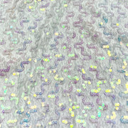 A close-up of a textured gray fabric covered in rows of iridescent sequins that form wavy, zigzag patterns. The sequins reflect a mix of pastel colors including purple, yellow, green, and blue.