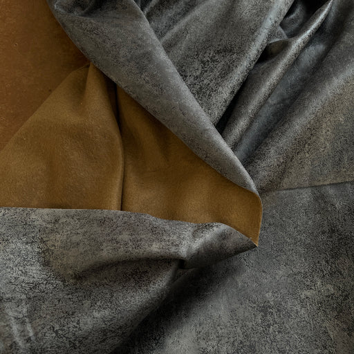A close-up of draped fabric showing two material/colorways: a mottled charcoal gray exterior and a smooth mustard-brown interior, arranged on a warm brown surface.