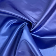 A close-up of shiny royal blue satin or silk-like fabric arranged in soft folds and swirls, filling the frame.
