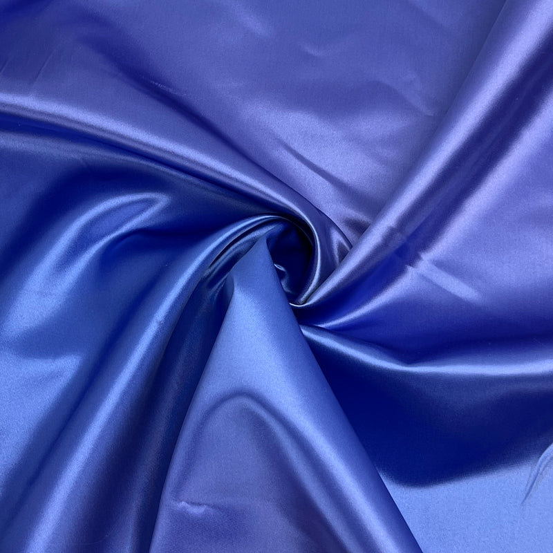 A close-up of shiny royal blue satin or silk-like fabric arranged in soft folds and swirls, filling the frame.