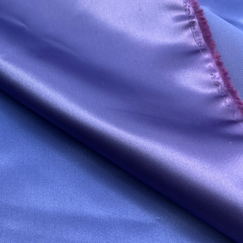 A close-up of a purple satin fabric with a diagonal fold showing its smooth, lustrous texture and a stitched frayed edge.