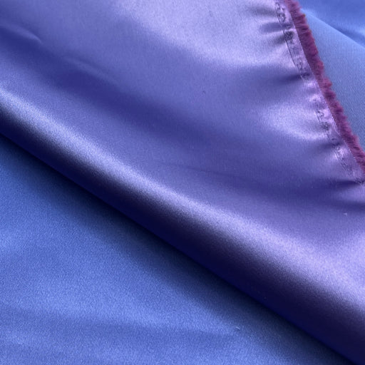 A close-up of a purple satin fabric with a diagonal fold showing its smooth, lustrous texture and a stitched frayed edge.