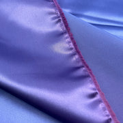 A close-up of shiny lavender-purple satin fabric with a visible magenta-pink stitched edge and fringe.