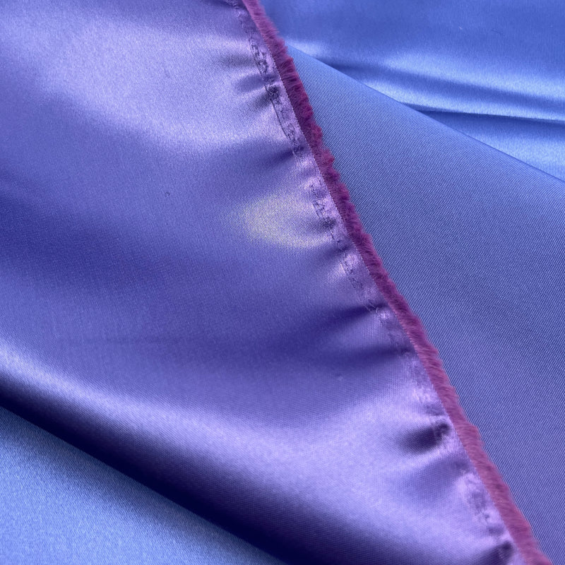 A close-up of shiny lavender-purple satin fabric with a visible magenta-pink stitched edge and fringe.