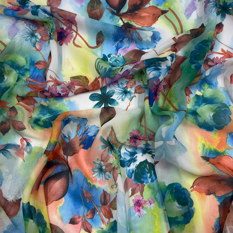 A close-up of lightweight draped fabric featuring a vibrant watercolor floral print with blue, green, pink, orange, and brown tones on a pale background.