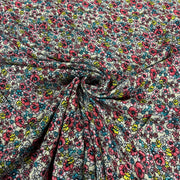 A close-up of a lightweight fabric with a dense, all-over floral print. The pattern features pink and red flowers, teal and blue blossoms, and small yellow accents on a white background, styled with a central swirl to show the material's drape and texture.